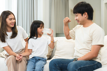 Happy Asian family, mother and father cheering little 6 year old daughter drinking milk from glass, raised arm muscles and fist for good health, sitting on sofa at home. Selective focus on child