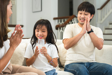 Happy Asian family, mother and father clapping hands cheering their little 6 year old daughter drinking milk from glass with smiling face in morning, sitting on sofa at home. Selective focus on child