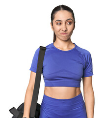 Athletic Caucasian young woman with gym bag on studio background confused, feels doubtful and...