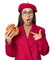 Chic Caucasian woman eating croissant in red coat and beret on studio background pointing to the side
