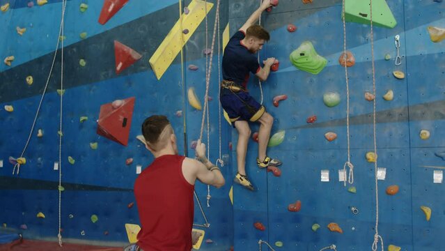 Man climbing wall in gym while his workout partner with prothetic leg handling rope and belays him