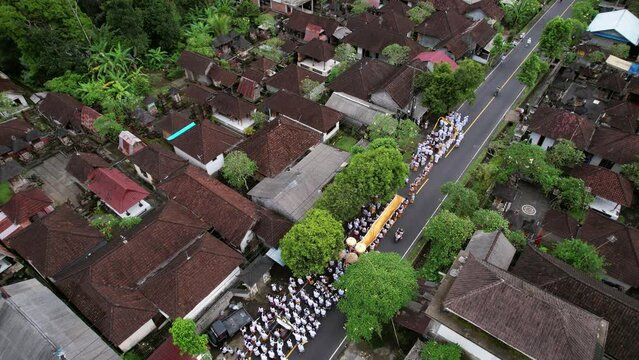 Village street is blocked off to one side by a ceremonial procession, many people in white clothes walking together. Various religious events are celebrated on the island of Bali