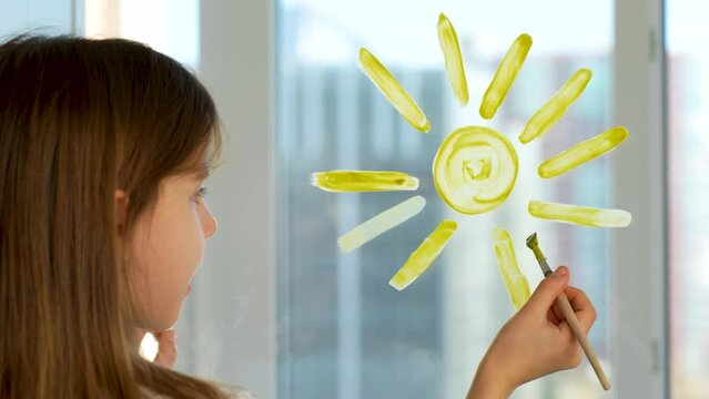 Little girl drawing sun with paintbrush and yellow paint on glass surface