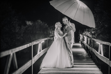 Valmiera, Latvia - August 13, 2023 - A bride and groom stand under an umbrella on a bridge at night, in a tender pose, captured in a black and white photograph.