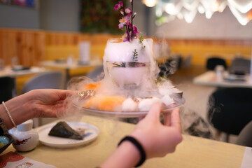 A hand holds a very exotic plate with various types of sushi and sashimi against a warm and...