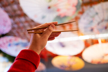 A hand holds two chopsticks for eating Asian food against a warm and colorful Japanese restaurant...