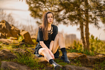  girl with a calm face sits on rocks in a black dress in the park