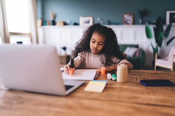 Engaged African American girl, 6-8 years old, diligently drawing at a wooden table, with a laptop, smartphone, and stationery around her. She's wearing a soft beige sweater, concentrating on studying