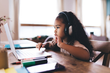 Adorable African American little girl sitting at wooden table typing on laptop and listening to...