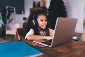 Adorable African American little girl sitting at wooden table typing on laptop and listening to...