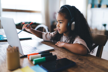 Adorable African American little girl sitting at wooden table on laptop computer and listening...