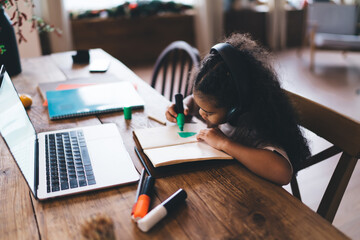 Concentrated African American girl, approximately 6-8 years old, wearing headphones while sitting at a wooden table with colorful school supplies, laptop computer during distance online class studying
