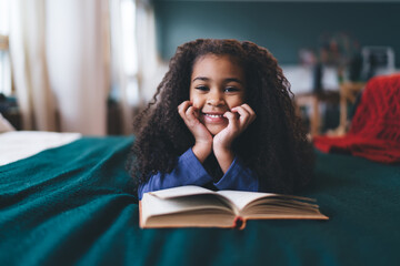 Joyful African American girl, aged 6-8, lying on her stomach on a green blanket, hands on cheeks, with a book open in front of her. Her bright smile and sparkling eyes, paired with a blue top - obrazy, fototapety, plakaty