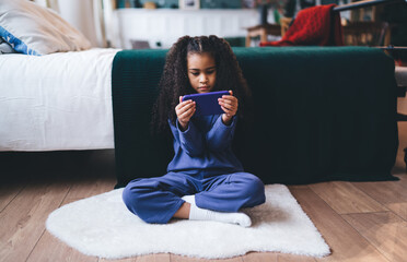 Focused African American girl, 6-8 years old, sitting cross-legged on a fluffy white rug, deeply...