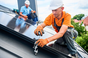 Workers building photovoltaic solar panel system on rooftop of house. Men technicians in helmets...