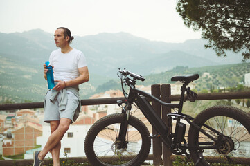 Athletic man holding a bottle of water, dreamily looking way, relaxing after biking in mountains,...
