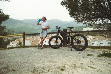 Obraz premium Handsome muscular man cyclist drinks water from sports bottle, standing by wooden fence next to electric mountain bike, on rural road overlooking white houses with red tile roofs in medieval village