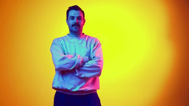 Young man in pink tie-dye sweatshirt posing with crossed and arms in pockets with calm expression on gradient yellow orange background in neon. Concept of human emotions, casual fashion, lifestyle