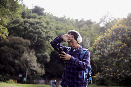 Young Asian man in a casual outfit, sporting a beanie, plaid shirt, and headphones, checks his phone with a puzzled expression on his morning nature walk. A backpack rests comfortably on his shoulder.