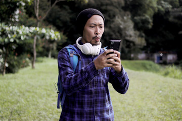 Asian man in a casual outfit, sporting a beanie, plaid shirt, and headphones, looks at his phone with a surprised expression on his morning nature walk. A backpack rests comfortably on his shoulder