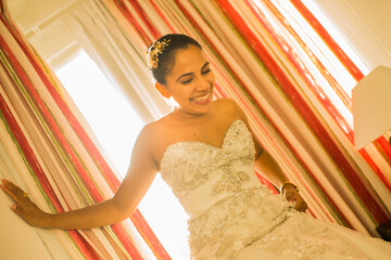 mujer joven latina sonriendo mientras usa su vestido de bodas y baja unas escaleras  - obrazy, fototapety, plakaty