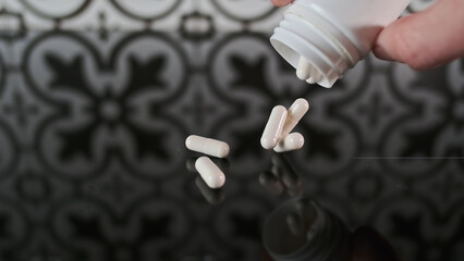 Hand pours white pill capsules from a container onto a glass reflective surface. Antidepressant...