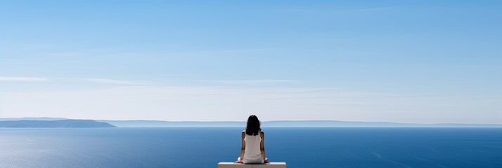 A woman is sitting on the edge facing the sea. The view is peaceful and serene, with vast expanses of water stretching out in the distance - obrazy, fototapety, plakaty