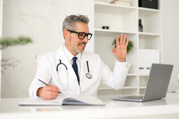 Cheerful senior doctor engages virtually with patients or colleagues, using hand gestures to explain or communicate during an online consultation or meeting from his office. Contemporary telemedicine
