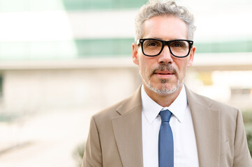 A senior businessman in glasses with grey hair poses confidently outdoors, his sharp beige suit and...