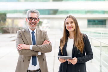 A poised senior executive with grey hair stands confidently with his arms crossed, accompanied by a female professional holding a tablet, symbolizing leadership and expertise.