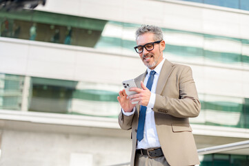 A grey-haired business executive beams as he looks at his smartphone after receiving good news. The lines of the office building in the background suggest a professional yet contemporary work setting.