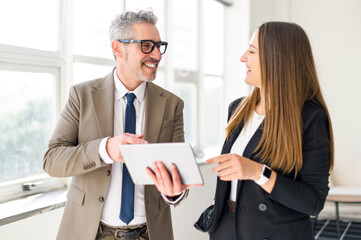 Senior businessman with grey hair, laughing and sharing ideas with a young female professional while looking at a tablet, in a well-lit modern office. Image represents positive aspects of mentorship
