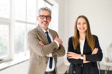 A charismatic senior entrepreneur with grey hair stands arms crossed next to a young professional woman, both smiling confidently in a modern office, reflecting a successful business partnership
