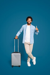 A cheerful young Brazilian man with a captivating smile holds a silver suitcase, giving a thumbs-up against a vibrant blue background, exuding a sense of excitement and anticipation for travel