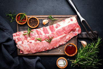 Raw pork ribs with rosemary, spices and barbecue sauce on oak wooden cutting board prepared for cooking on black kitchen table background, top view - 779782175