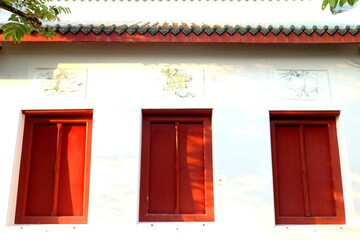 Close painted red wood windows row on white cement wall of building in Buddism temple and ancient native Thai art above with some shadow