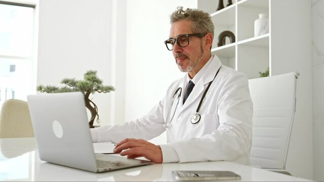 A genial mature doctor in a white coat waves hello into laptop during online appointment, seated in a well-organized clinic office that reflects a clean and efficient medical environment, taking notes