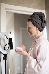 Woman using eye patches, smiling and holding cosmetic cream jar, looking at smartphone. Blogger recording skincare routine, vlogging