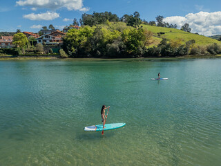hispanic girls couple paddleboarding on river past village houses on summer day
