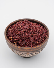 A round brown bowl with decorative carving filled with Sumac. The bowl is set against a plain white background