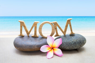 Mom wooden font on stone with plumeria flower over tropical beach background, mother's day card background idea