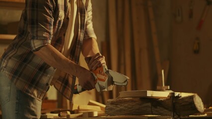 Male woodworker working in garage. Man professional carpenter working with wooden materials in workshop sawing wood using cordless mini chainsaw. - obrazy, fototapety, plakaty