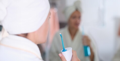 Young woman in gown and towel on head and brushing her teeth with irrigator in front of mirror in bathroom closeup. Dental care concept