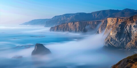 The ocean is calm and the sky is blue. The mist is rising from the water and the rocks are in the foreground