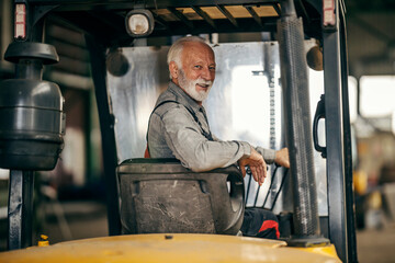 A senior factory worker is driving forklift and looking over his shoulder at the camera.
