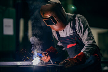 A metallurgy worker with helmet on head is welding in heavy industry plant.