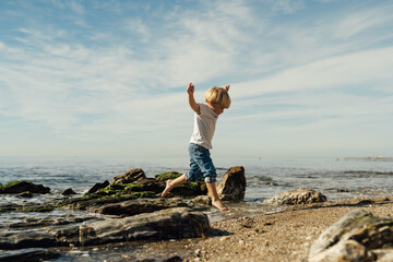 little blond boy playing on the seashore. child playing on the seashore. boy stands on stones in...