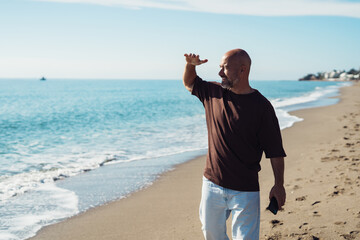 A mature adult man, wearing a T-shirt and jeans, happily walks along the seashore. A smiling man walks along the beach on a sunny day. A bald man with a beard walks along the seashore.