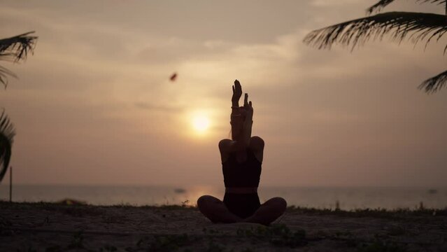 Woman practices yoga on ocean at sunset. Silhouette sporty yoga girl doing yoga pose asana exercise meditation at sunset on beach in nature close up. Health morning routine, harmony lifestyle concept.