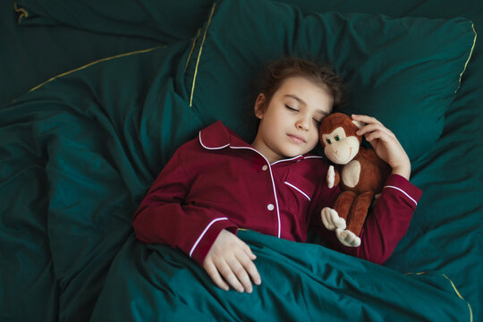 7-year-old girl in red pajamas sleeps on green bed with plush monkey toy. Baby looks peaceful and content as she sleeps.